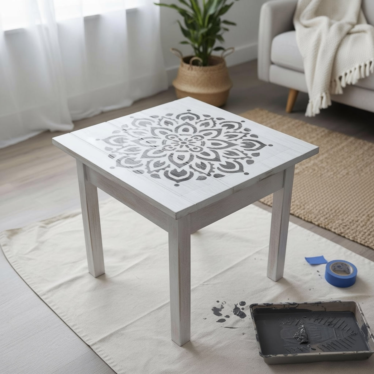 a white wooden table with a stencil design on it, placed on a white cloth on a wooden floor. There is a blue painter's tape and a tray of paint on the floor next to the table. In the background, there is a couch and a potted plant.