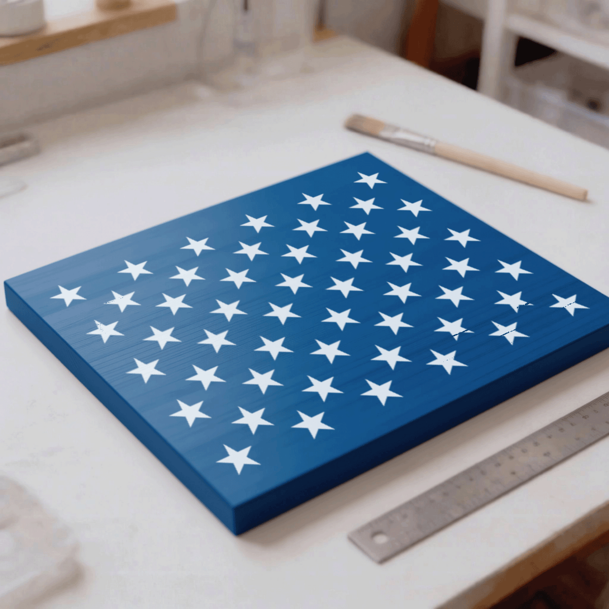 a blue square canvas with white stars on it, placed on a white surface. There is a paintbrush and a ruler nearby, suggesting that the canvas is being prepared for painting or measuring.