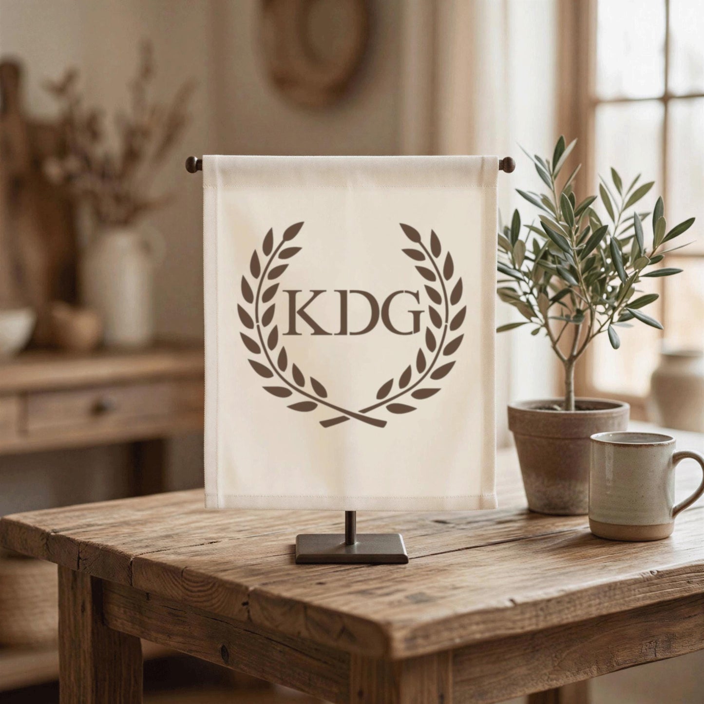 a wooden table with a white banner displaying the initials "KDG" in a wreath design, accompanied by a potted plant and a coffee mug.