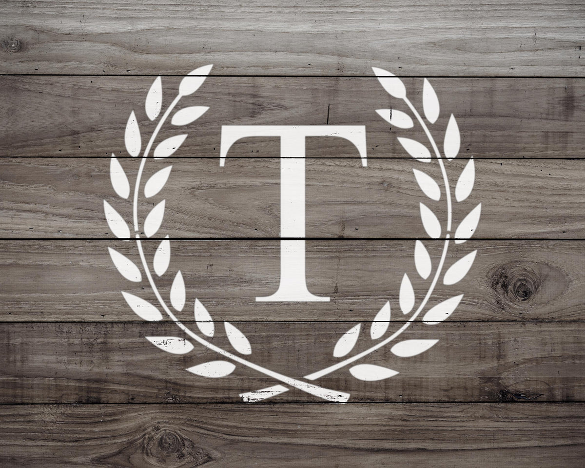 A monogrammed letter 'T' in the center of a laurel wreath design on a wooden background.
