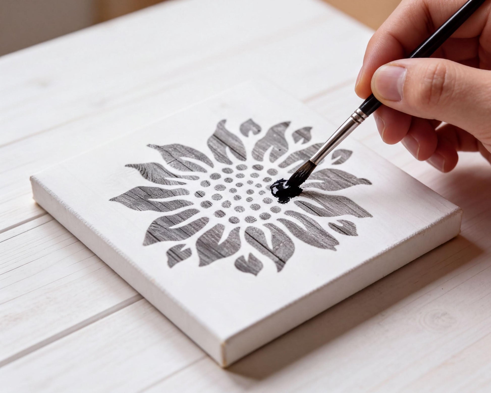 A hand is painting a stencil of a flower on a white canvas.