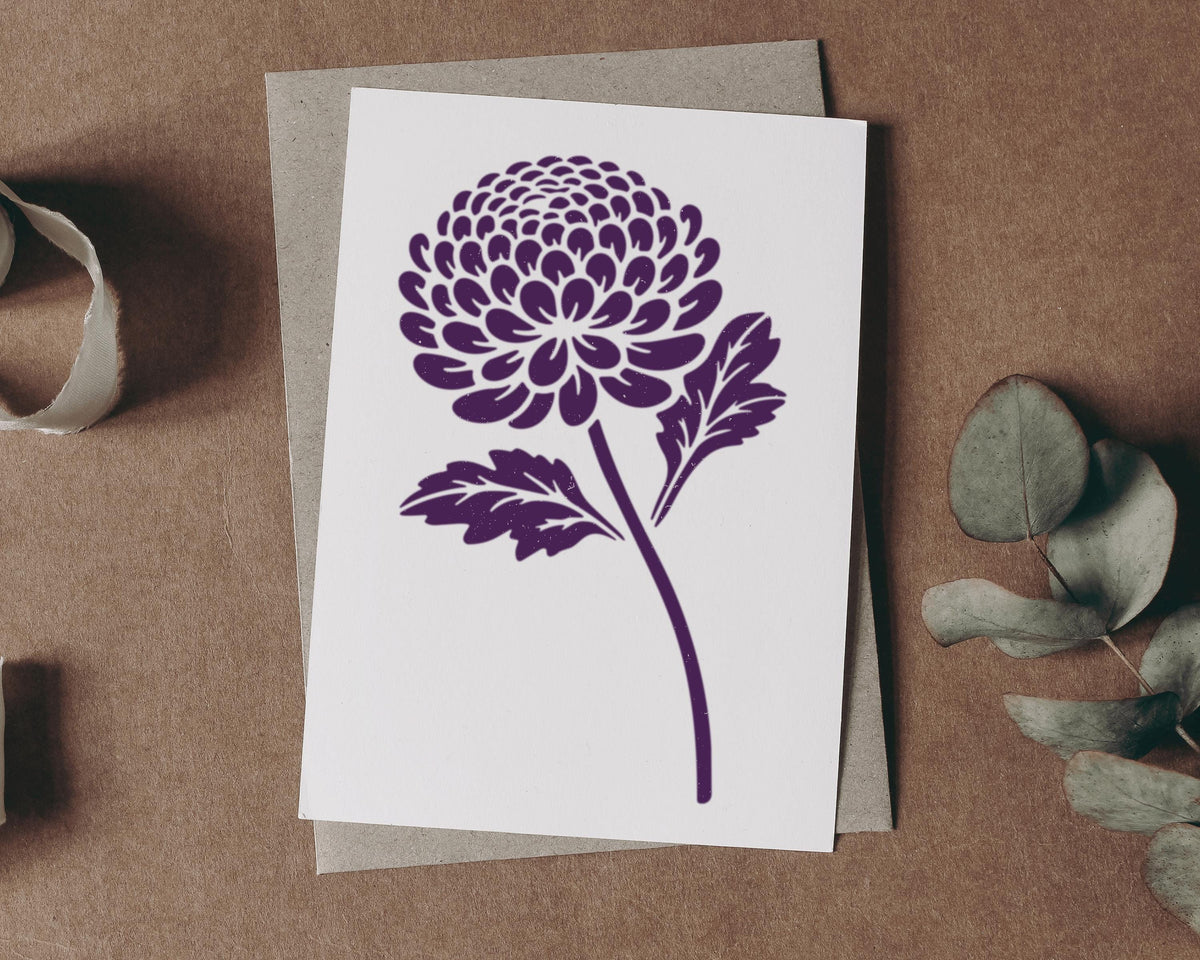 a white card with a purple flower design on it, placed on a brown surface. There are also some greenery and a roll of tape in the background.
