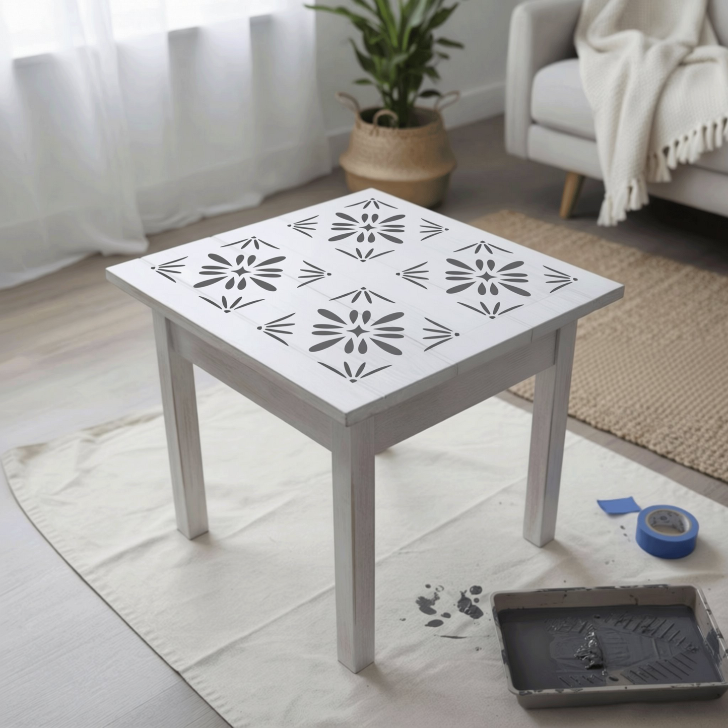 a white table with a stencil design on its surface, placed on a white cloth on a wooden floor. There is a potted plant in the background, and a couch can be seen in the background as well.