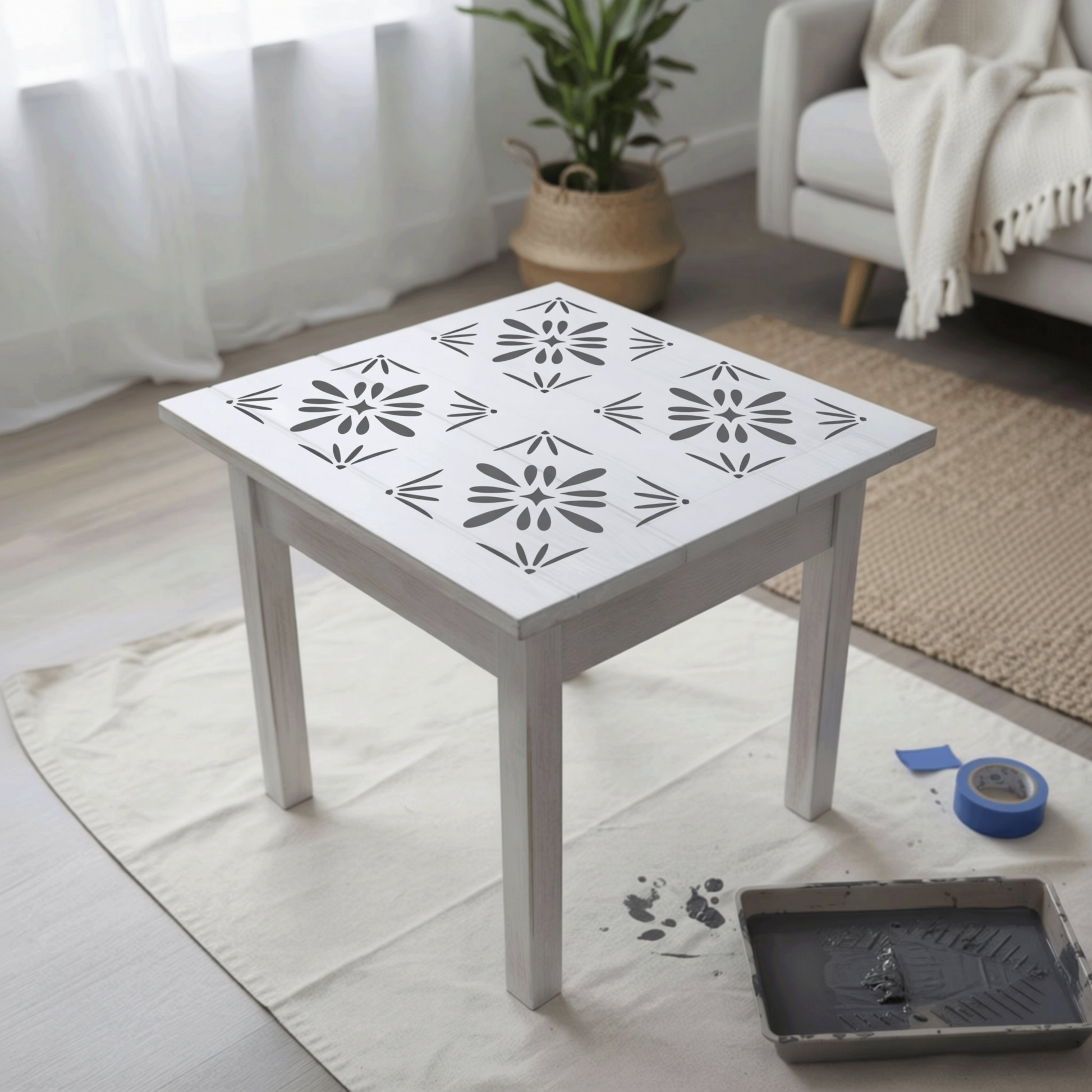 a white table with a stencil design on its surface, placed on a white cloth on a wooden floor. There is a potted plant in the background, and a couch can be seen in the background as well.