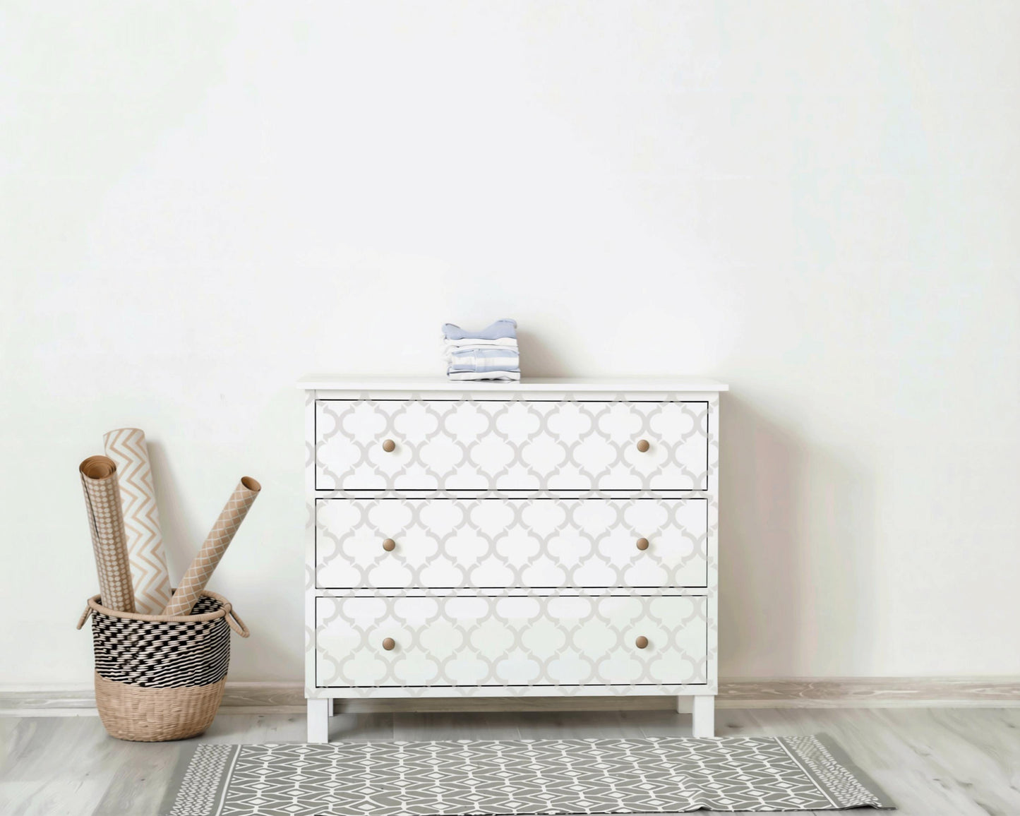 a white three- drawer dresser with a geometric pattern, placed against a white wall. Next to the dresser is a woven basket filled with rolls of wrapping paper.