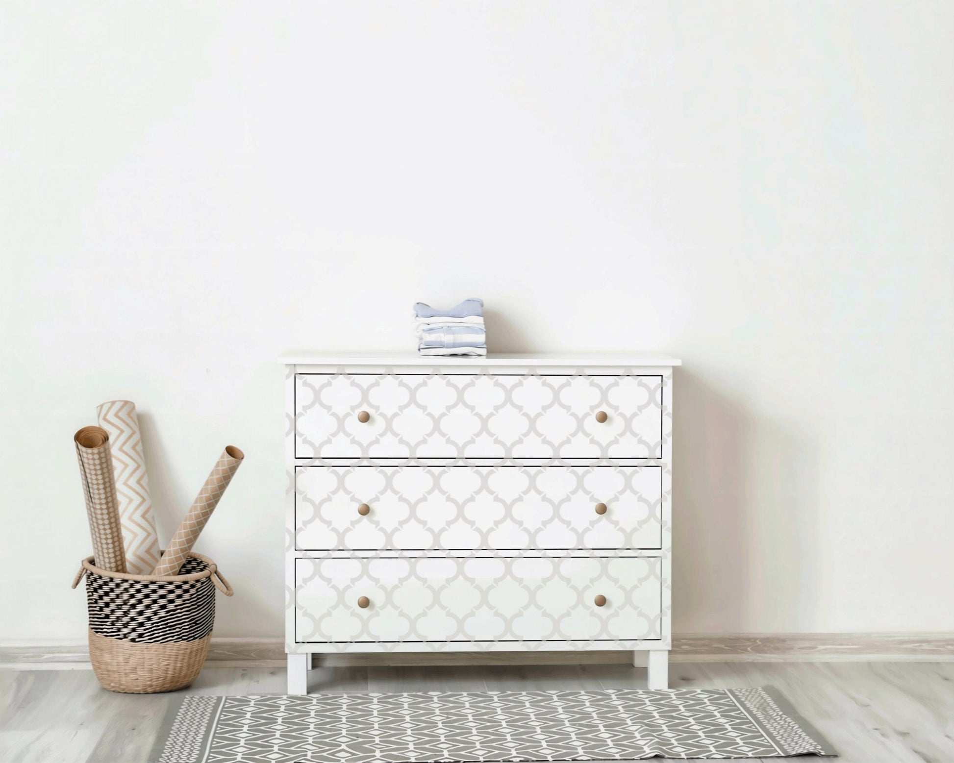 a white three- drawer dresser with a geometric pattern, placed against a white wall. Next to the dresser is a woven basket filled with rolls of wrapping paper.