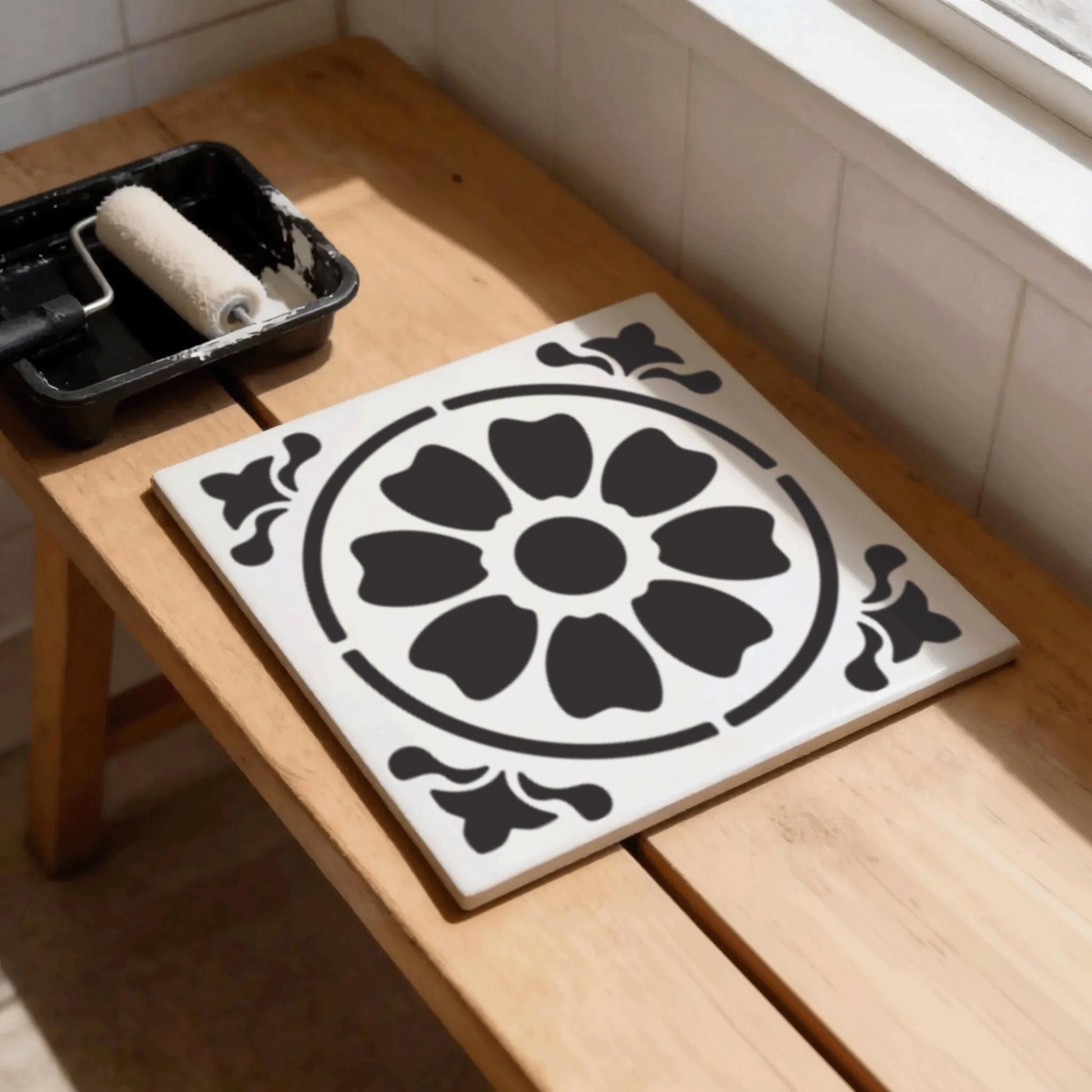 a wooden bench with a white tile featuring a black floral design, and a paint roller with some paint on it, suggesting that the bench is being prepared for painting or decoration.