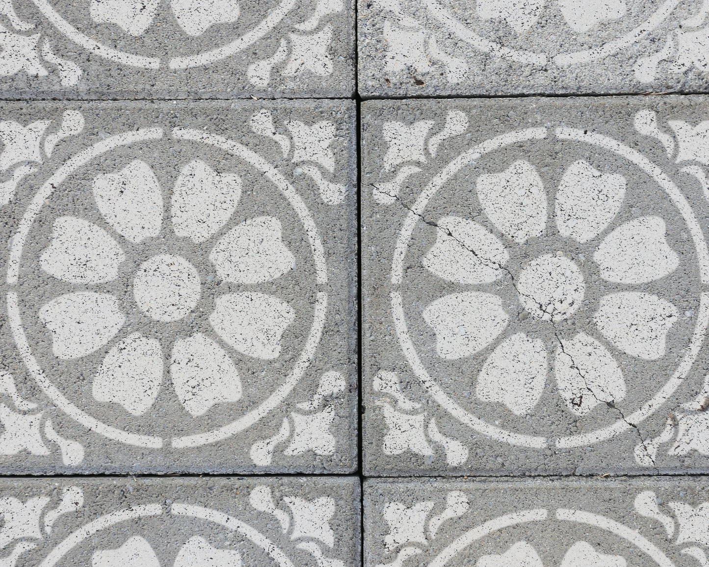 a close-up view of a patterned concrete surface with a repeating design of white flowers or petals. The pattern is symmetrical, with the flowers arranged in a circular or oval shape. The concrete surface appears to be made of small blocks or tiles, creating a textured and visually interesting surface.