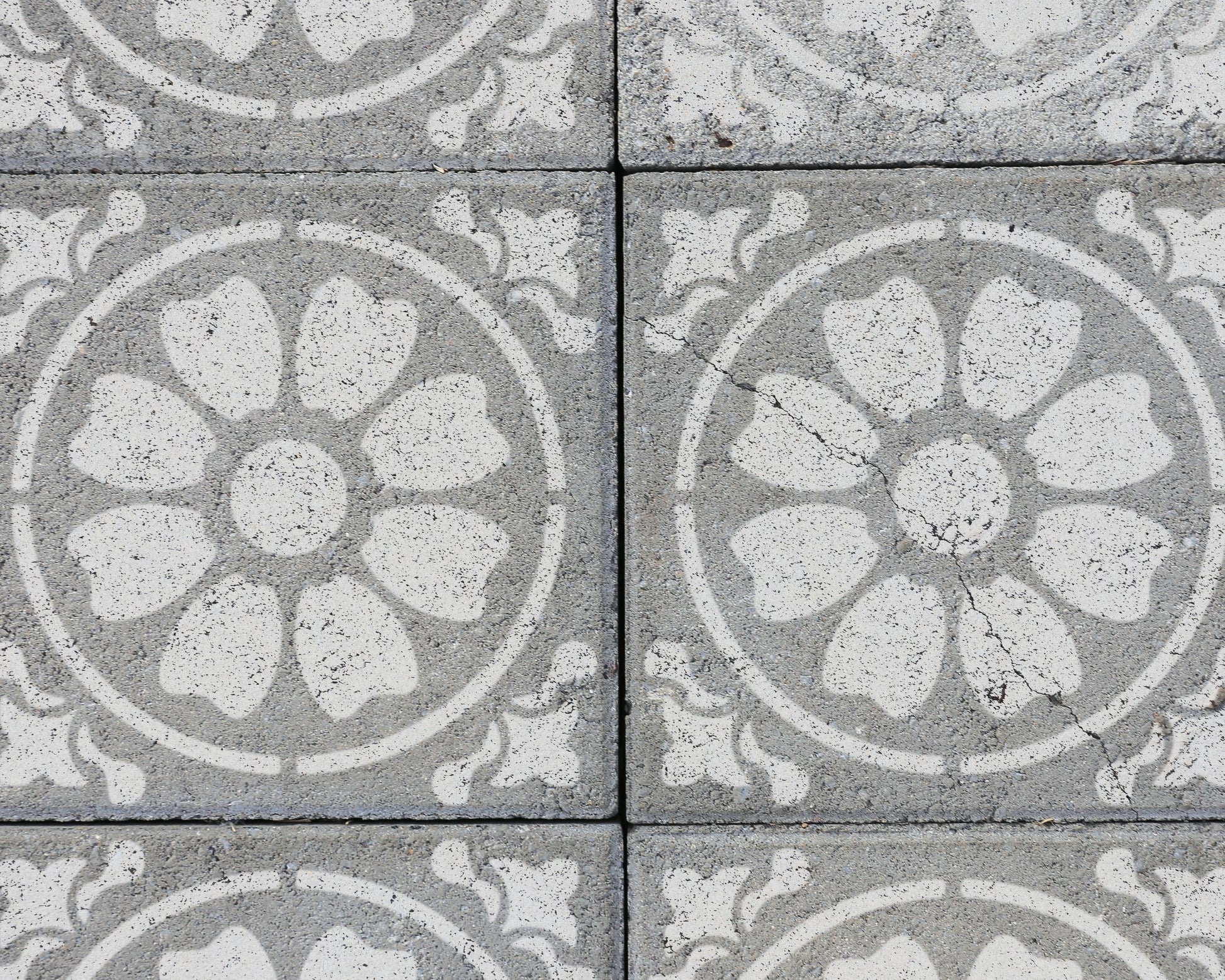 a close-up view of a patterned concrete surface with a repeating design of white flowers or petals. The pattern is symmetrical, with the flowers arranged in a circular or oval shape. The concrete surface appears to be made of small blocks or tiles, creating a textured and visually interesting surface.
