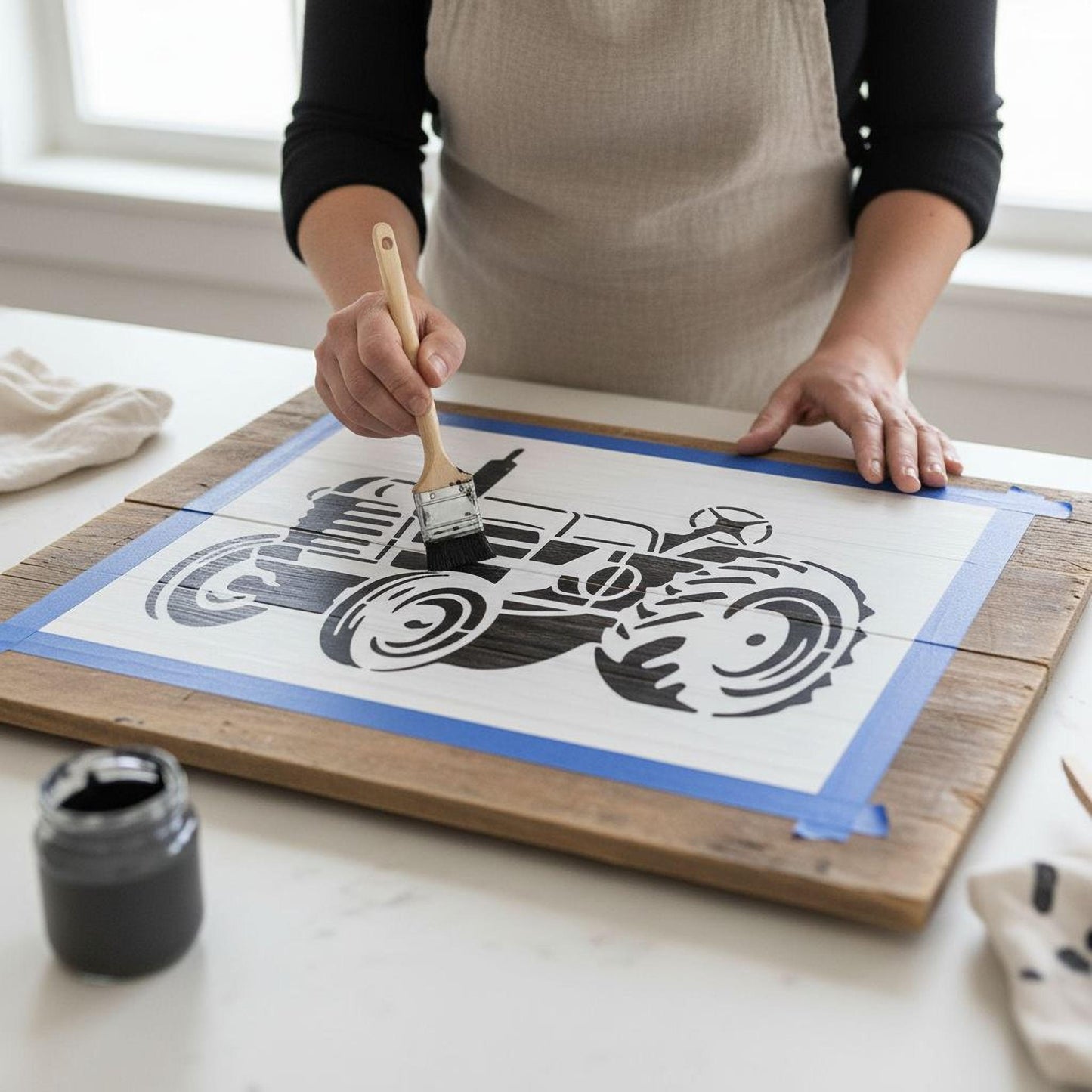 A person is painting a wooden board with a stencil of a tractor, using a brush to apply paint.