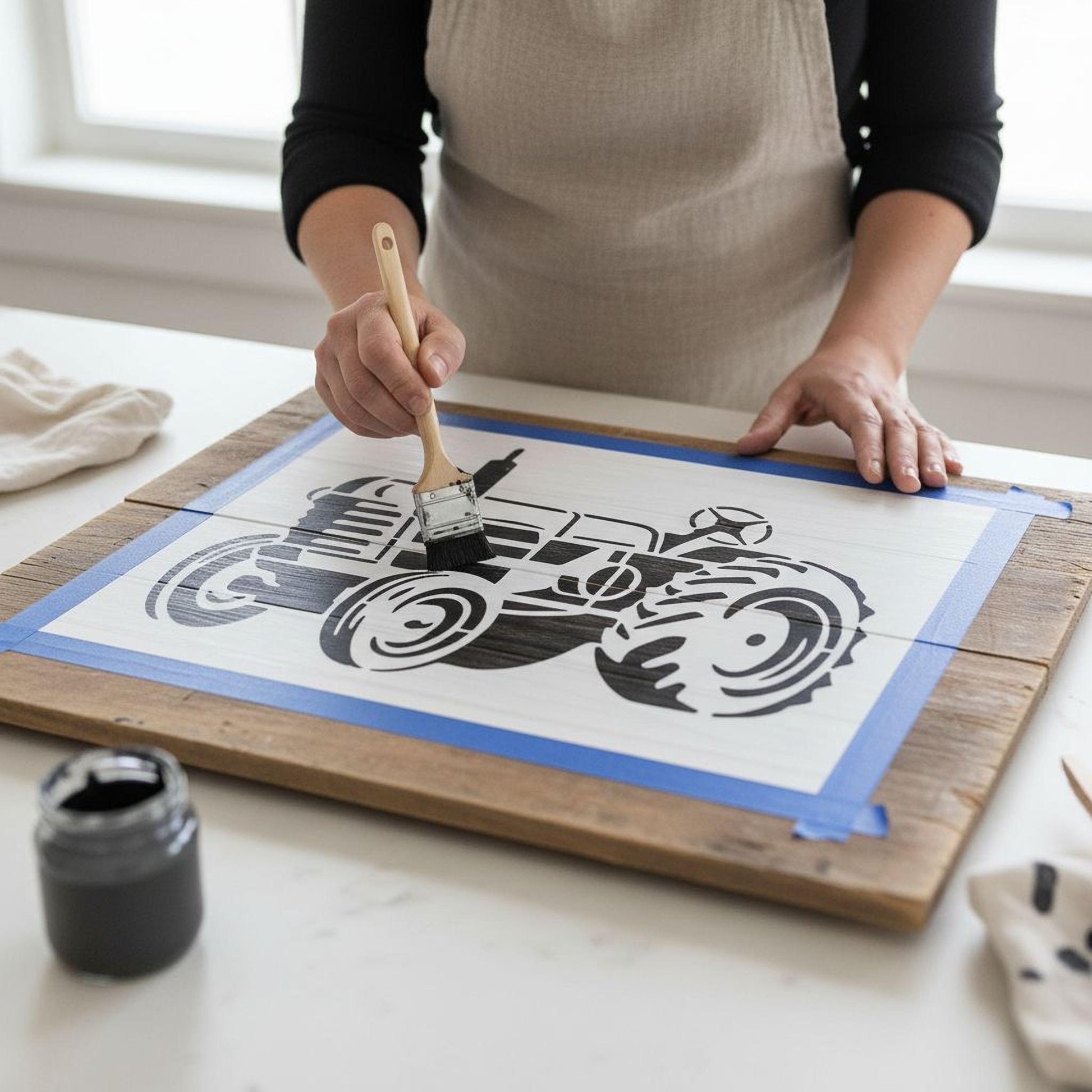 A person is painting a wooden board with a stencil of a tractor, using a brush to apply paint.