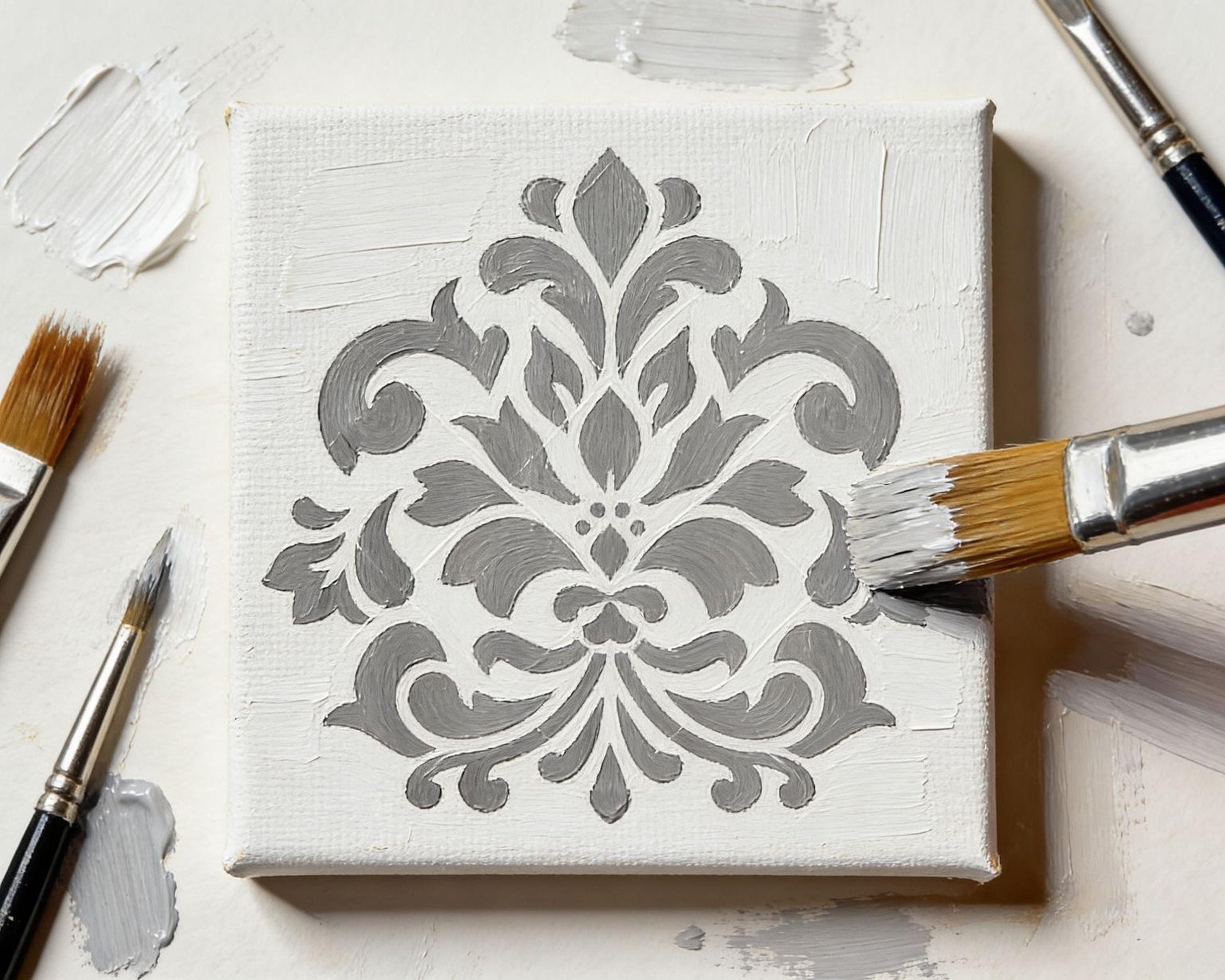 a white canvas with a decorative gray and white pattern painted on it, surrounded by various paintbrushes and paint splatters on a white surface.
