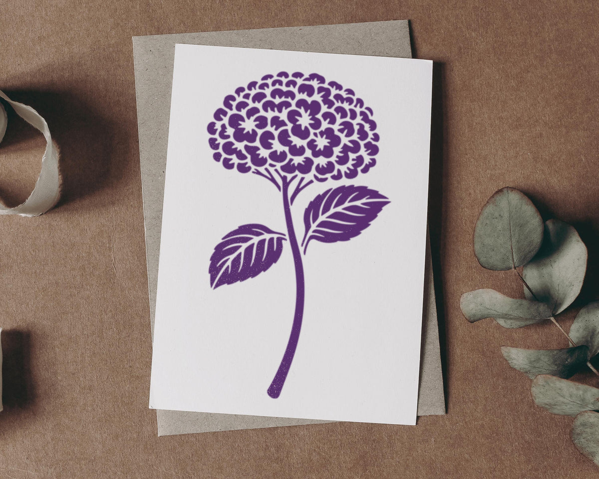 a white card with a purple floral design on it, placed on a brown surface next to some greenery.