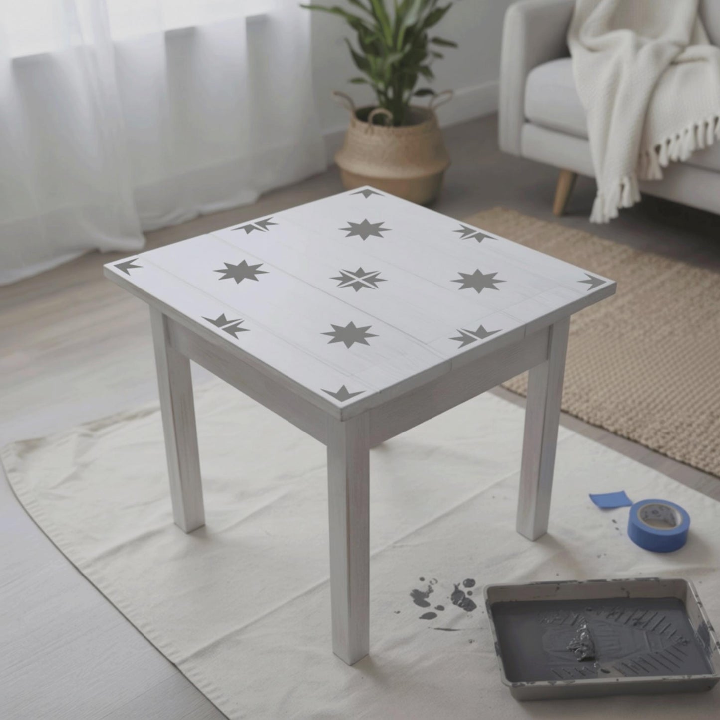 a white wooden table with a stencil design of stars on it, placed on a white cloth on a wooden floor. There is a blue painter's tape and a tray of paint on the floor next to the table. In the background, there is a couch and a potted plant.