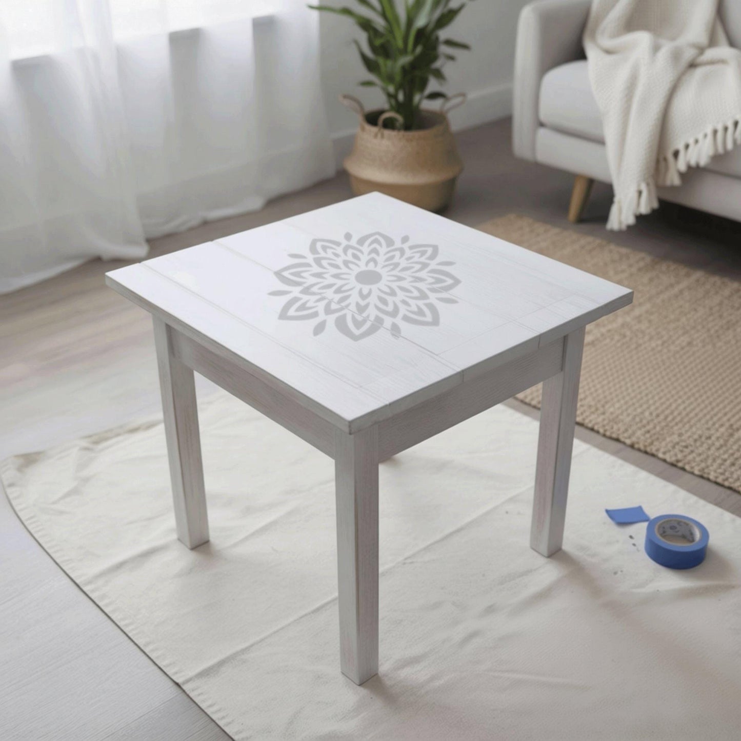 a white wooden table with a stencil design on its surface, placed on a white cloth on a wooden floor. In the background, there is a couch and a potted plant, creating a cozy and inviting atmosphere.