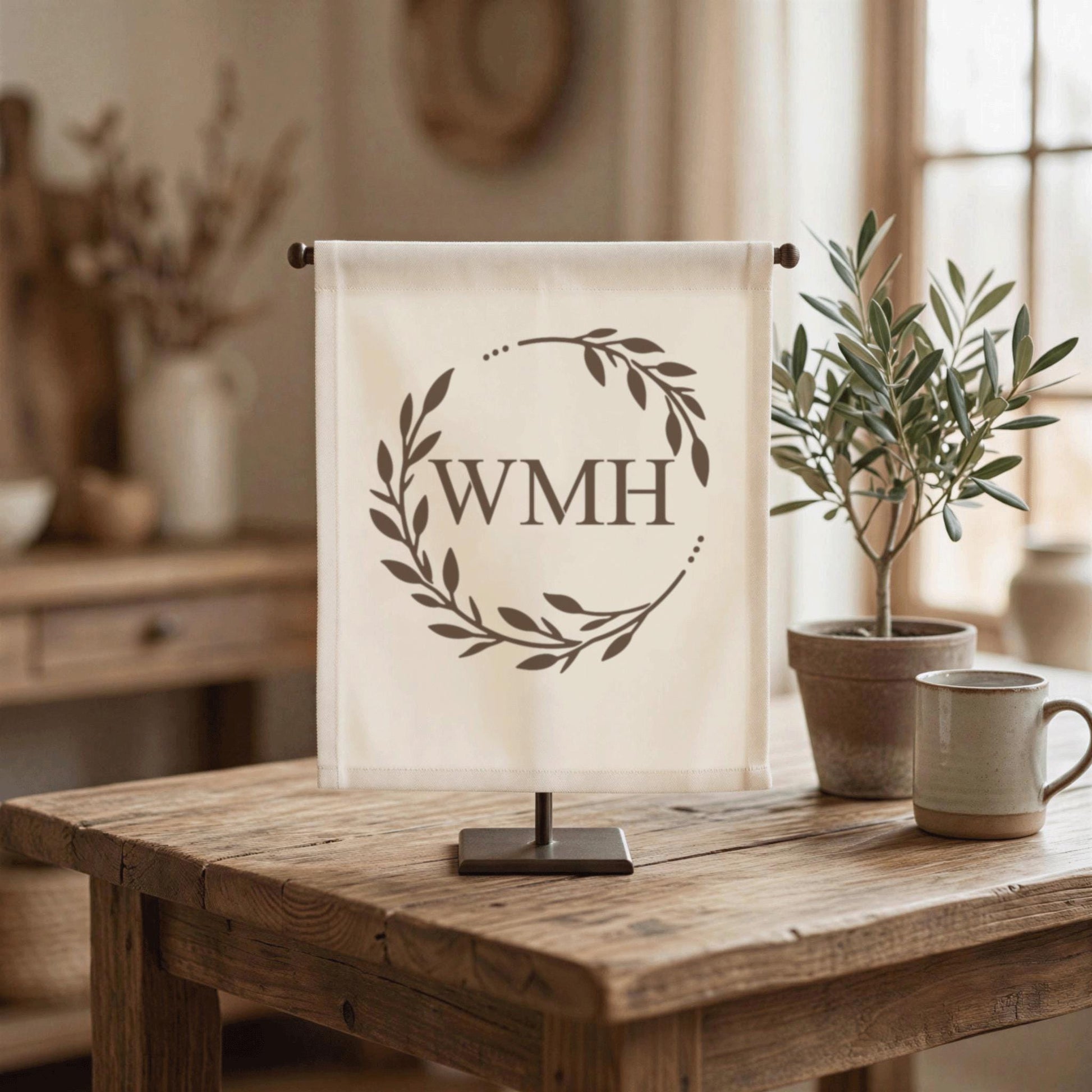 a wooden table with a white banner displaying the initials "WMH" in the center, surrounded by a wreath design. There are two potted plants on the table, one on the left and one on the right, along with a coffee cup and a vase.
