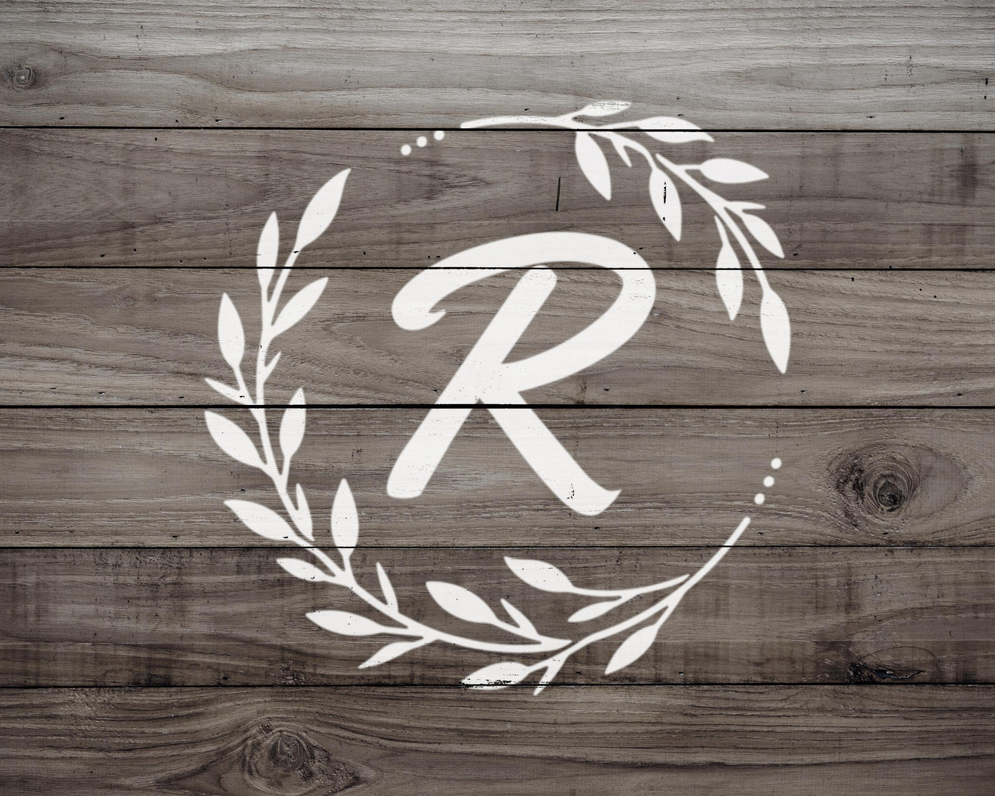 A monogrammed white letter "R" is displayed on a wooden background with a wreath of leaves surrounding it.