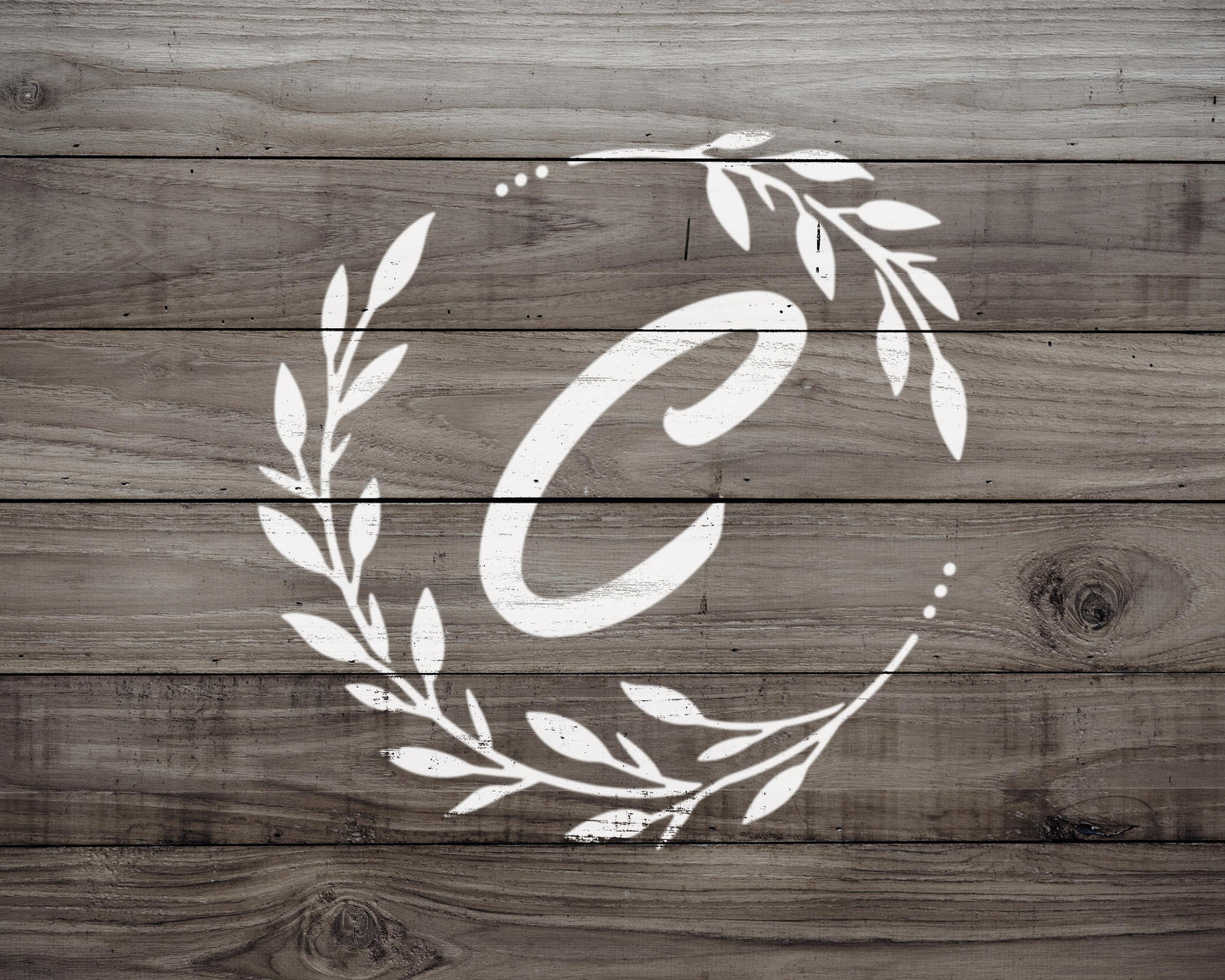 A monogrammed white letter 'C' with a wreath of leaves and branches surrounding it, set against a wooden background.