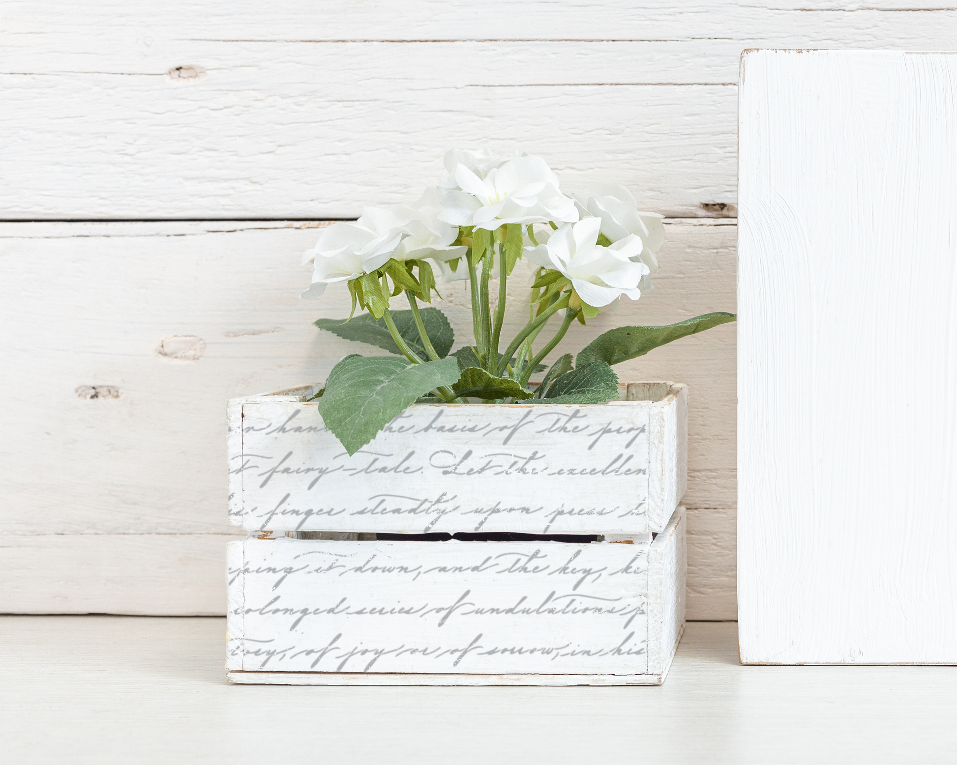 A white wooden crate with a decorative text pattern on it, containing a bouquet of white flowers. The crate is placed on a white wooden surface, with a white wooden board visible in the background.