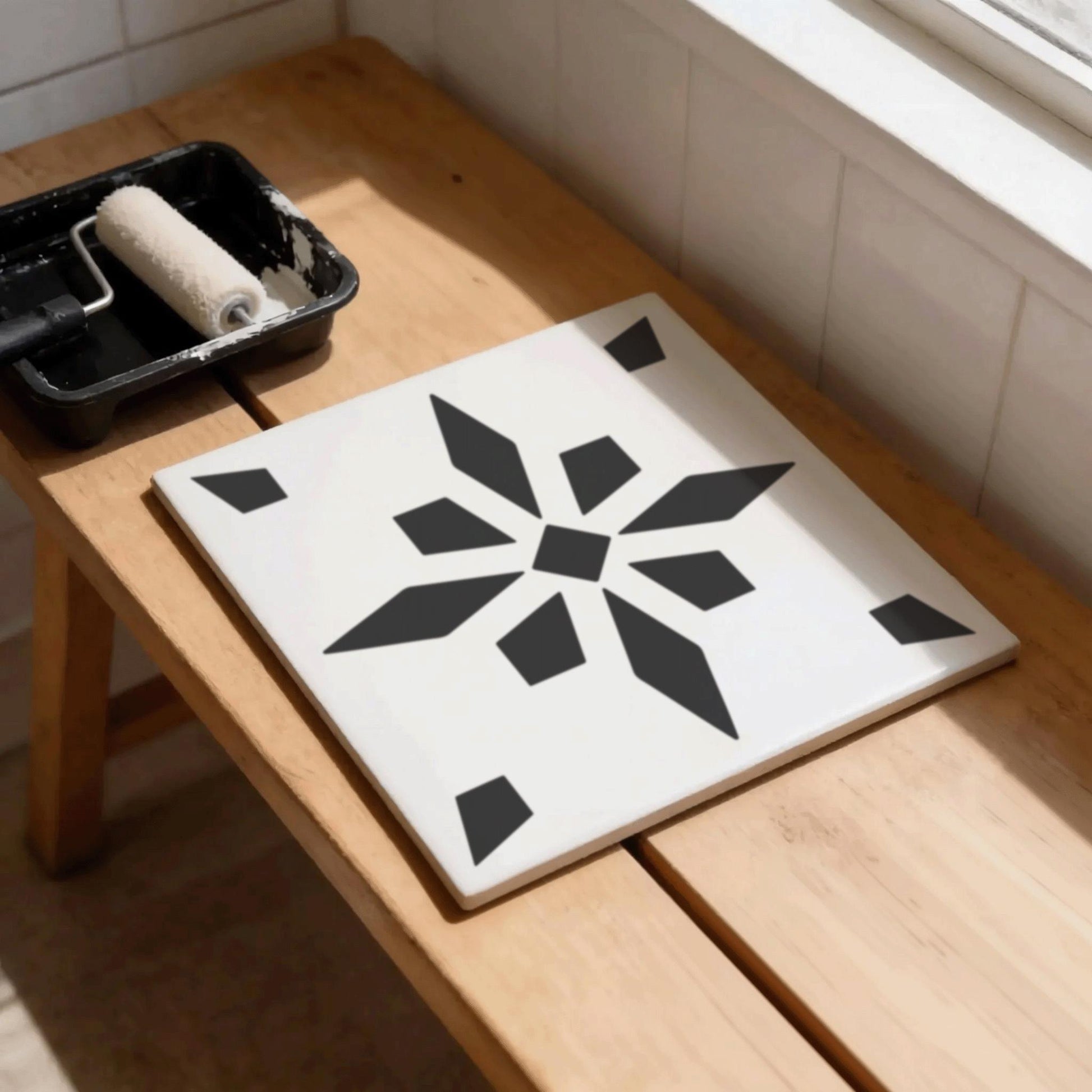 a wooden table with a white tile featuring a black geometric design, and a paint roller and tray of paint are placed on the table.