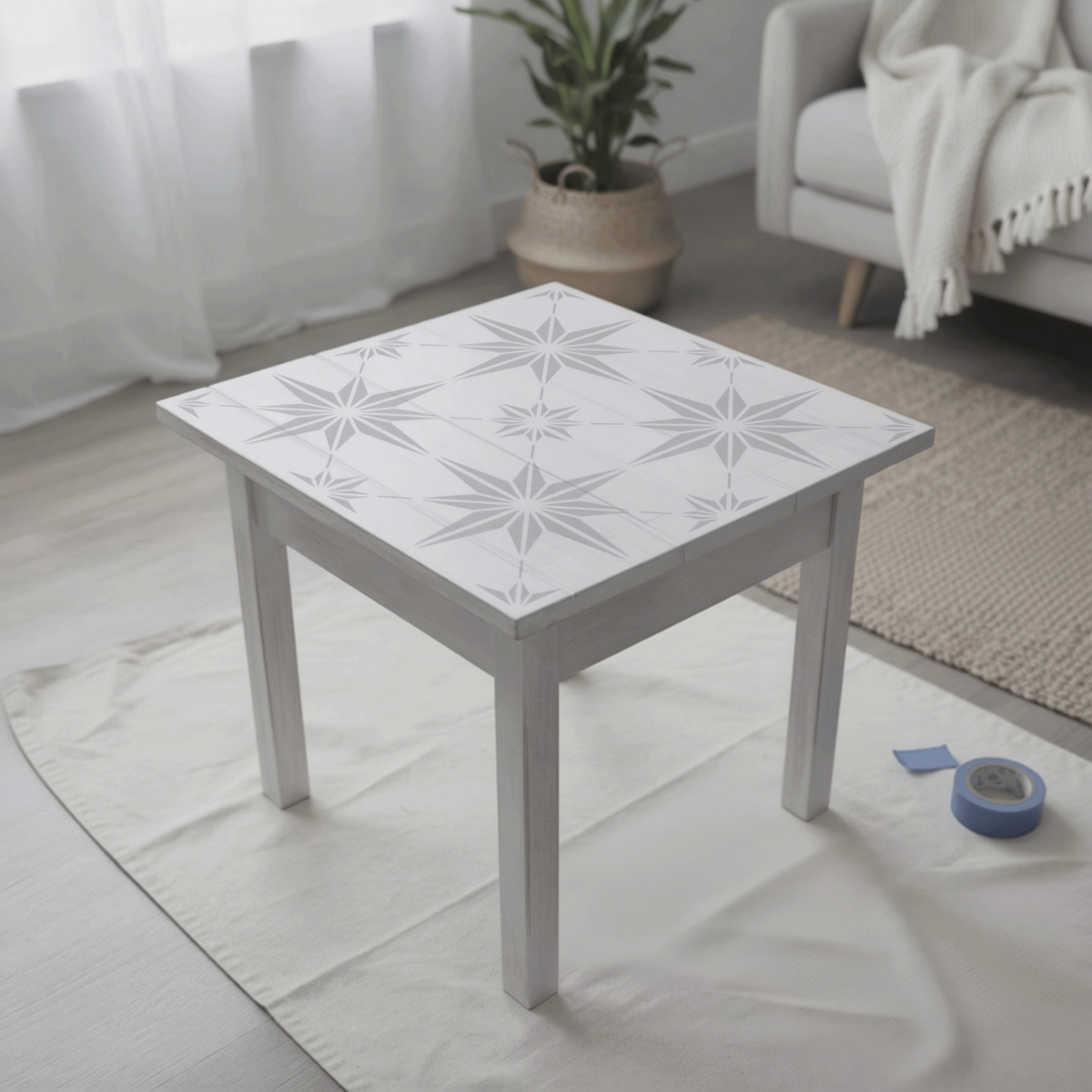 a white square table with a star-shaped design painted on its surface, placed on a white cloth on a wooden floor. In the background, there is a couch and a potted plant, creating a cozy and inviting atmosphere.