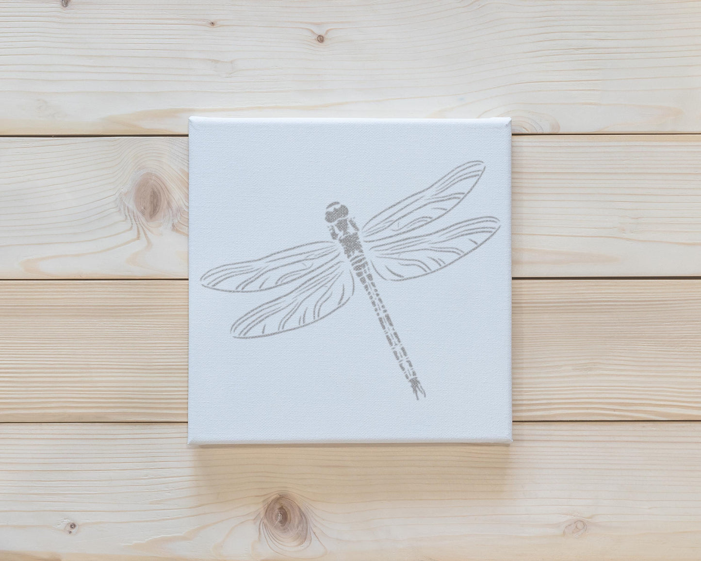 a white canvas with a black and white illustration of a dragonfly on it, placed on a wooden surface.