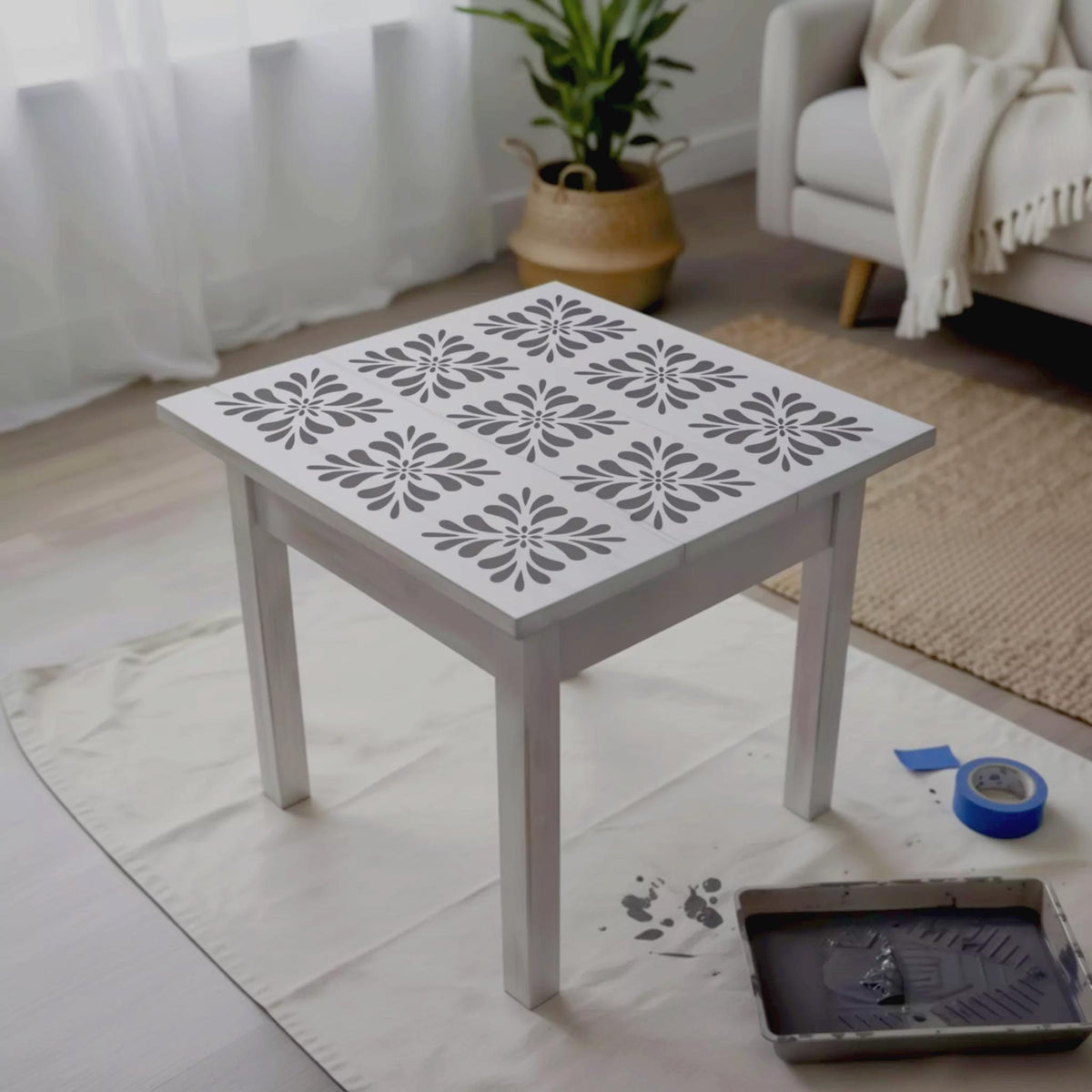 a white table with a stencil design on its surface, placed on a white cloth on a wooden floor. There is a potted plant in the background, and a couch can be seen in the background as well.
