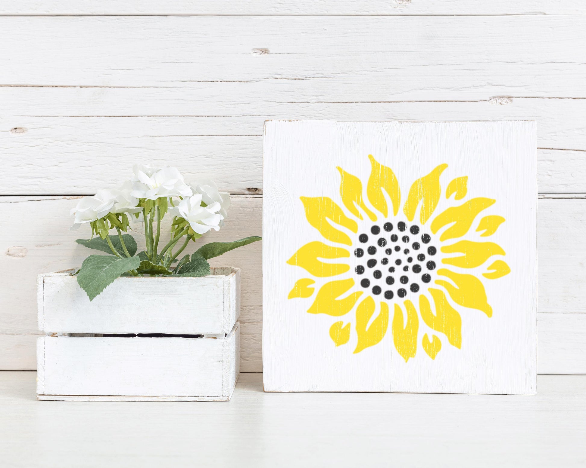 a white wooden crate with a potted plant inside, and a white square sign with a yellow sunflower design on it, placed on a white wooden surface.