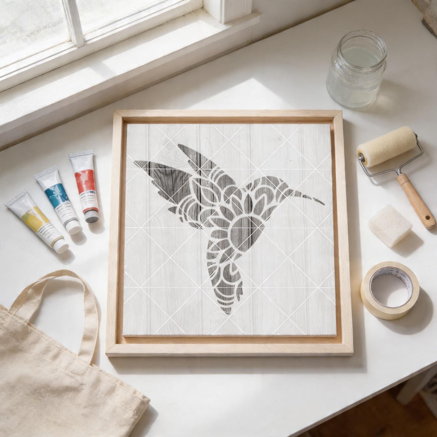 a wooden frame with a stencil of a bird, a roller brush, and various art supplies arranged on a white surface.