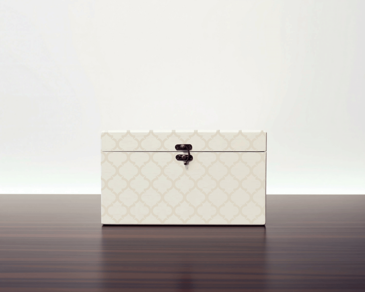 A white, patterned box with a black latch is sitting on a dark wooden surface.